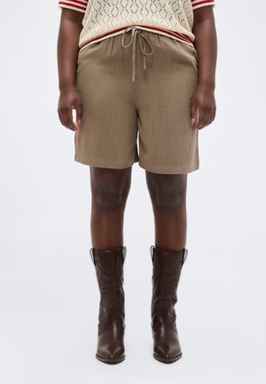 Personne portant un short beige à cordon, un haut en maille crème avec des rayures rouges et des bottes en cuir marron foncé mi-mollet, debout devant un fond blanc.