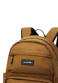 Ruskea kangasreppu mustilla yksityiskohdilla, etutaskulla, pehmustetuilla hihnoilla ja logopatchilla. Tekstuuri antaa kestävältä näyttävän vaikutelman.