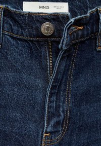 Jean en denim bleu foncé avec un bouton métallique et une fermeture éclair, avec des surpiqûres orange et une étiquette de marque blanche indiquant la taille.