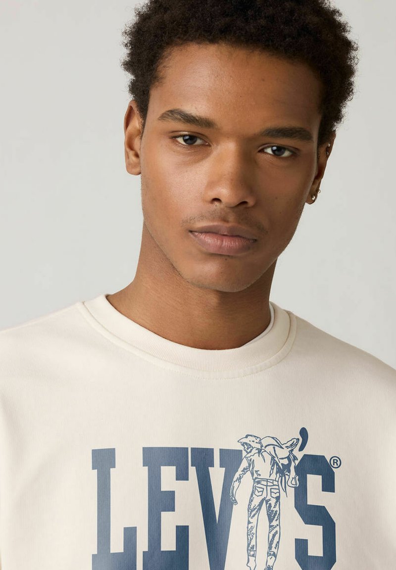 Sweat-shirt beige naturel avec le logo "LEVI'S" en bleu foncé, accompagné d'un graphisme représentant un homme portant un fagot. Col rond et texture douce.