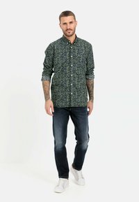 Blumenhemd in Marineblau mit gelben und grünen Akzenten, kombiniert mit dunklen Jeans und weißen Sneakers.