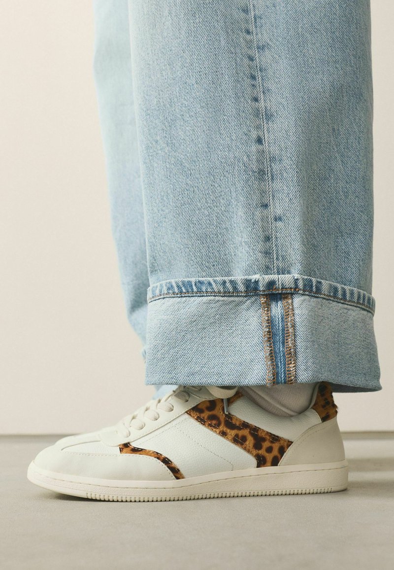 White leather sneaker with leopard print accents, flat sole, and tonal laces, paired with light blue cuffed denim jeans.