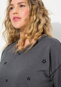 Sudadera de algodón gris con escote en forma de v, adornada con bordado de estrellas negras, costuras visibles y un diseño de corte relajado, con tela ligeramente texturizada.