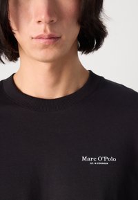 Black crewneck sweatshirt made of cotton, featuring a small white logo text on the left chest area. Soft texture, ribbed neckline.