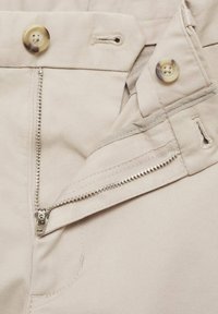 Pantalon en coton beige avec une fermeture éclair en métal, un bouton et des poches latérales. Le tissu a une texture lisse avec des détails de couture discrets.