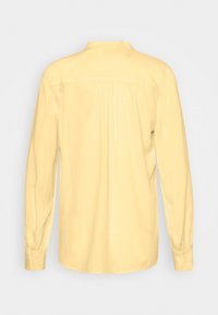 Camicia gialla a maniche lunghe vista da dietro, con polsini con bottoni e dettaglio arricciato sulla parte superiore della schiena.