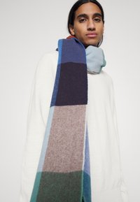 Écharpe en laine avec un patchwork géométrique dans des nuances de bleu, marron et vert, d'une texture douce et de forme rectangulaire.