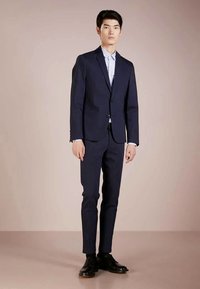 Fato azul-escuro com um corte ajustado, casaco de botão único, lapelas cortadas e calças slim. Combinado com uma camisa azul-clara e sapatos pretos.