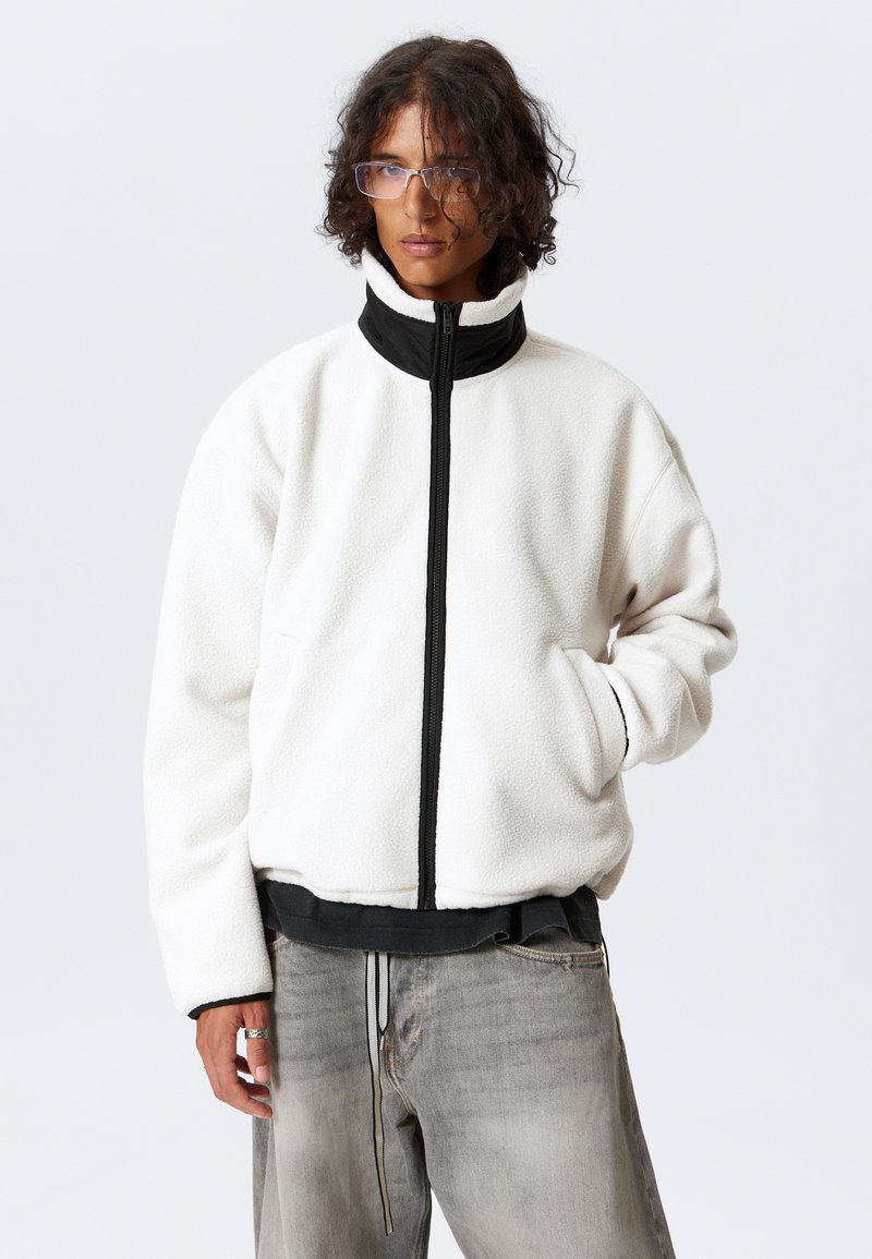 Chaqueta de forro polar blanca con un alto cuello negro, cierre con cremallera en el frente y bolsillos laterales. Textura suave; corte holgado; se lleva sobre una camiseta negra.