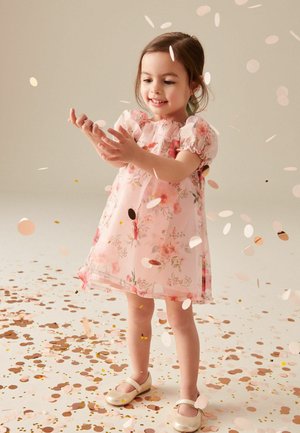 Robe florale rose avec des manches bouffantes, en tissu transparent et d'une coupe fluide. Enfant portant des chaussures dorées brillantes, entouré de confettis sur le sol.