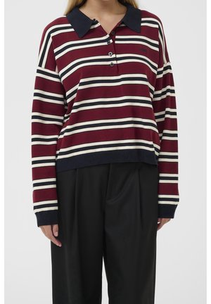 Personne portant un polo à manches longues avec des rayures horizontales bordeaux, blanches et noires, associé à un pantalon noir ample.