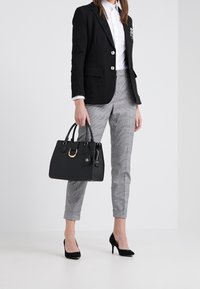 Black handbag with gold hardware, structured design, and brief handles. Model wears a black blazer, white shirt, and gray plaid trousers.