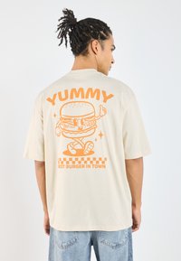 Beige oversized t-shirt featuring an orange cartoon burger graphic with the text "YUMMY" and "BEST BURGER IN TOWN" on the back.