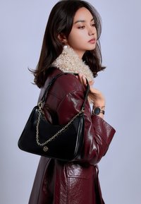 Vrouw in een bordeauxleren jas met een beige bontkraag, die een zwarte handtas met een gouden ketting vasthoudt, tegen een effen achtergrond.