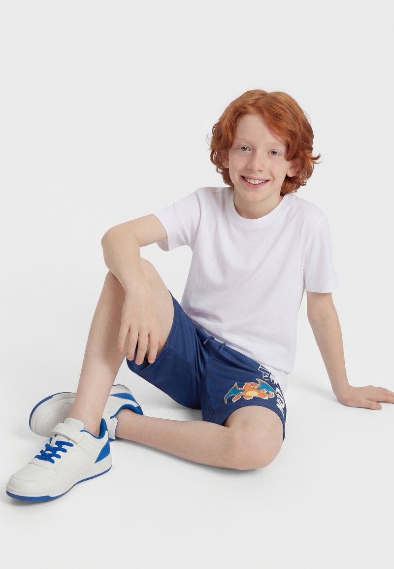 Κόκκινο-μαλλούχο αγόρι καθισμένο στο πάτωμα, φοράει λευκό μπλουζάκι, μπλε σορτς με σχέδιο καρτούν και λευκά αθλητικά παπούτσια με μπλε λεπτομέρειες, χαμογελά στην κάμερα.