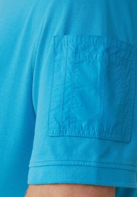 Turkusowa koszula z krótkim rękawem o fakturowanej tkaninie, z dużą kieszenią naszywaną na górnej części rękawa i prążkowanym ściągaczem na końcu rękawa.