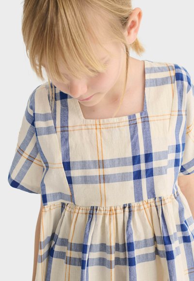 Enfant blonde portant une robe crème à carreaux bleus et oranges, regardant vers le bas, avec des manches courtes et un encolure carrée.