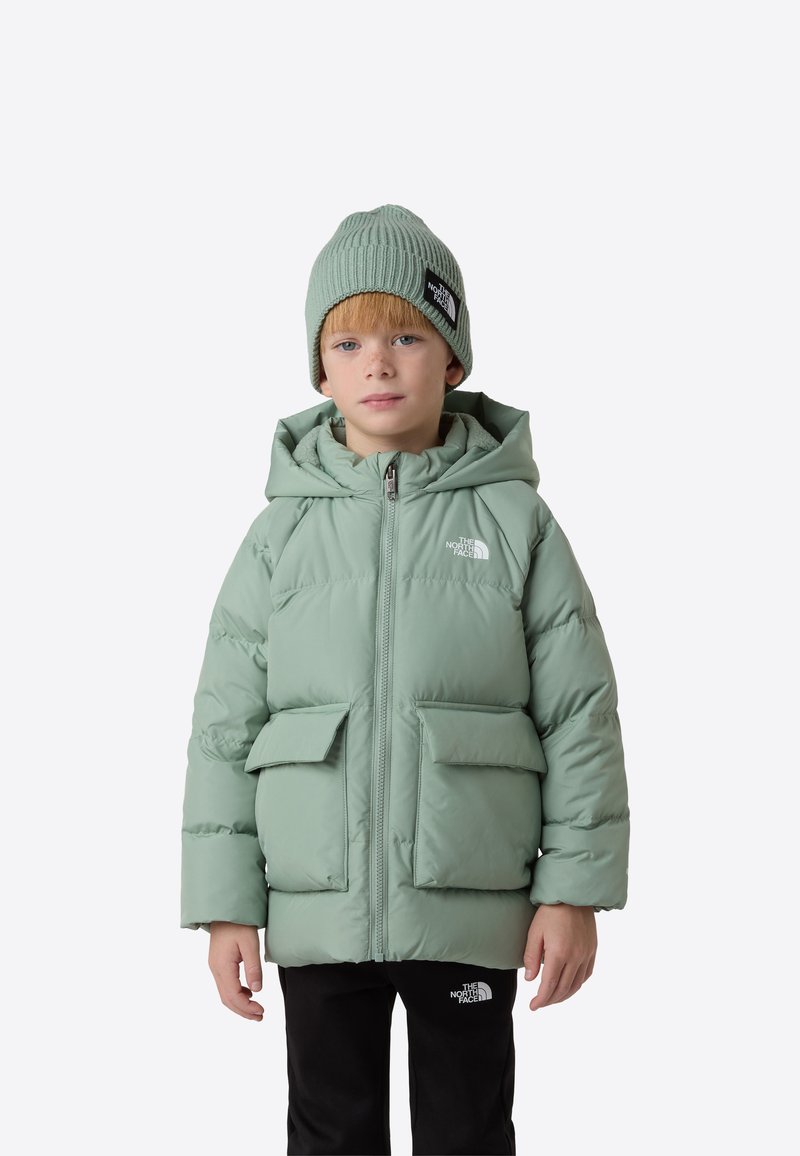 Grøn dunjakke med hætte og to forlommer, lavet af glat stof. Båret med en matchende strikket hue. Sorte bukser.