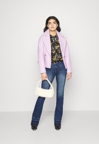 Casaco double-breasted em mistura de lã lavanda sobre uma camisa preta com padrão floral, combinado com jeans escuros de perna larga e uma bolsa trançada creme.