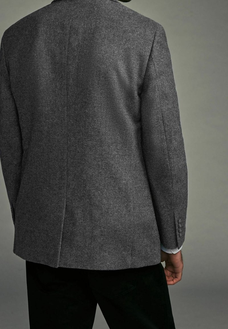 Homme portant un blazer gris à motif chevrons, vu de dos, avec un pantalon noir et un poignet de chemise clair visible.