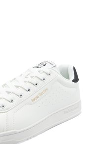 Weißer Ledersneaker mit perforierten Details, flachen Schnürsenkeln und einem schwarzen Fersenakzent. Logo in Gold auf der Seite und der Zunge aufgedruckt.