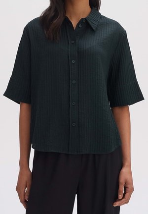 Nainen, jolla on musta, lyhythihainen, nappikiinnitteinen paita, jossa on poimutetun näköinen pinta ja terävä kaulus, yhdistettynä löysiin mustiin housuihin.