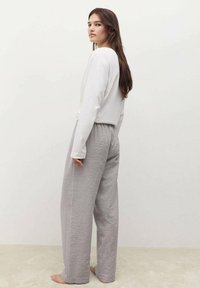 Jasnoszare i białe, paskowane szerokie spodnie z elastycznym pasem. Połączone z białą bluzką z długim rękawem, modelka stoi boso.
