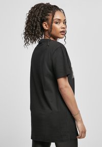 Černé oversized tričko s krátkými rukávy, kulatým výstřihem a hladkým povrchem. Má asymetrický lem a diskrétní logo na přední straně.