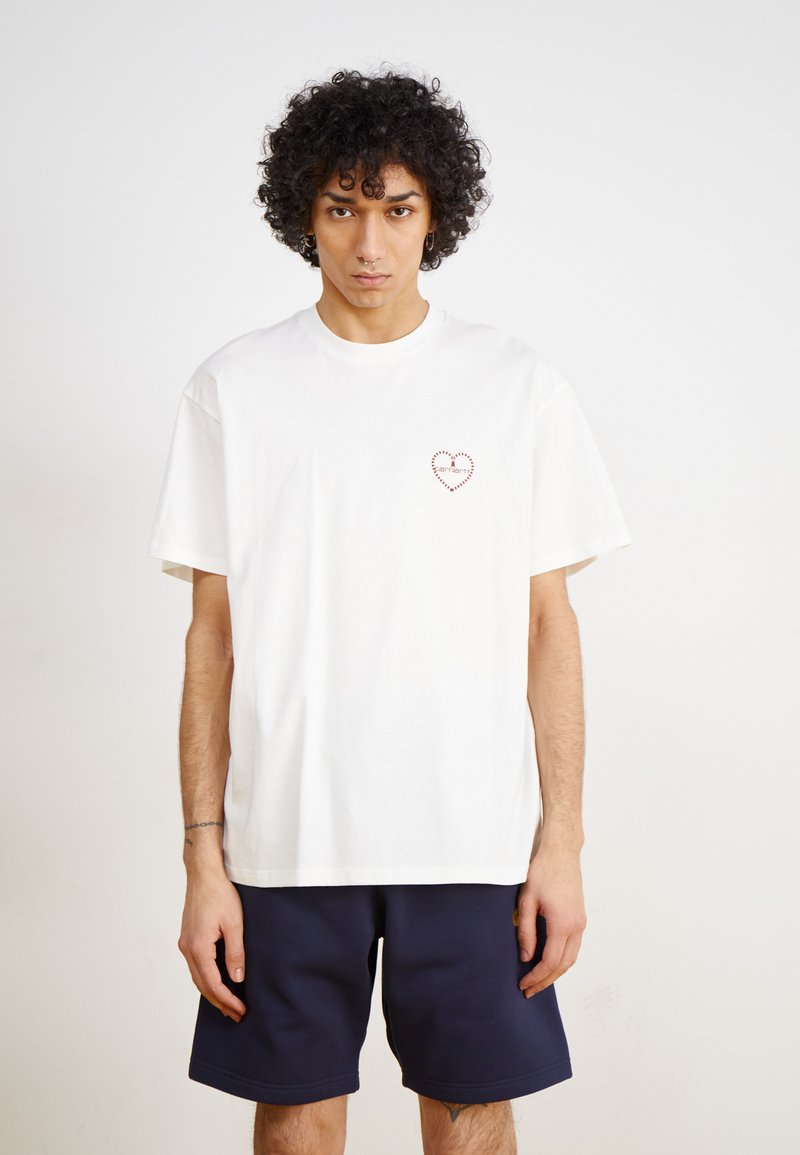 Witte katoenen t-shirt met een klein rood hartontwerp op de linkerkant van de borst, ontspannen pasvorm, gecombineerd met donkerblauwe shorts.