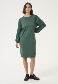 Robe en tricot verte avec des manches pleines et plissées, encolure ras du cou et poches latérales. Assortie avec des chaussures noires à enfiler. Texture lisse.