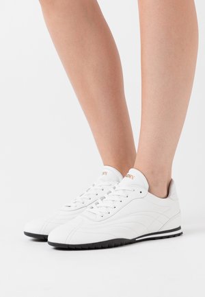 Benen met witte leren sneakers met zwarte zolen, witte veters en een subtiel gestikt ontwerp, staand op een wit oppervlak.