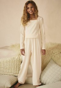 Light pink two-piece jumpsuit made of soft, ribbed fabric; features long sleeves, a square neckline, and relaxed fit pants.