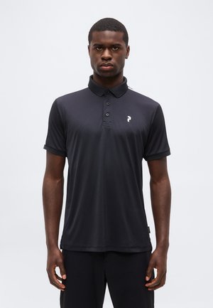 Hombre de pie mirando hacia adelante, vestido con una camiseta polo negra de manga corta con un pequeño logo blanco en el pecho izquierdo.