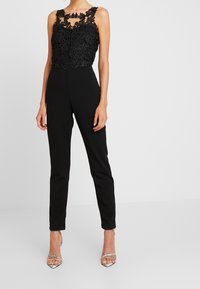 Mono negro con un corsé de encaje con patrones intrincados y detalle de ojal, pantalones ajustados y tacones plateados con tiras.