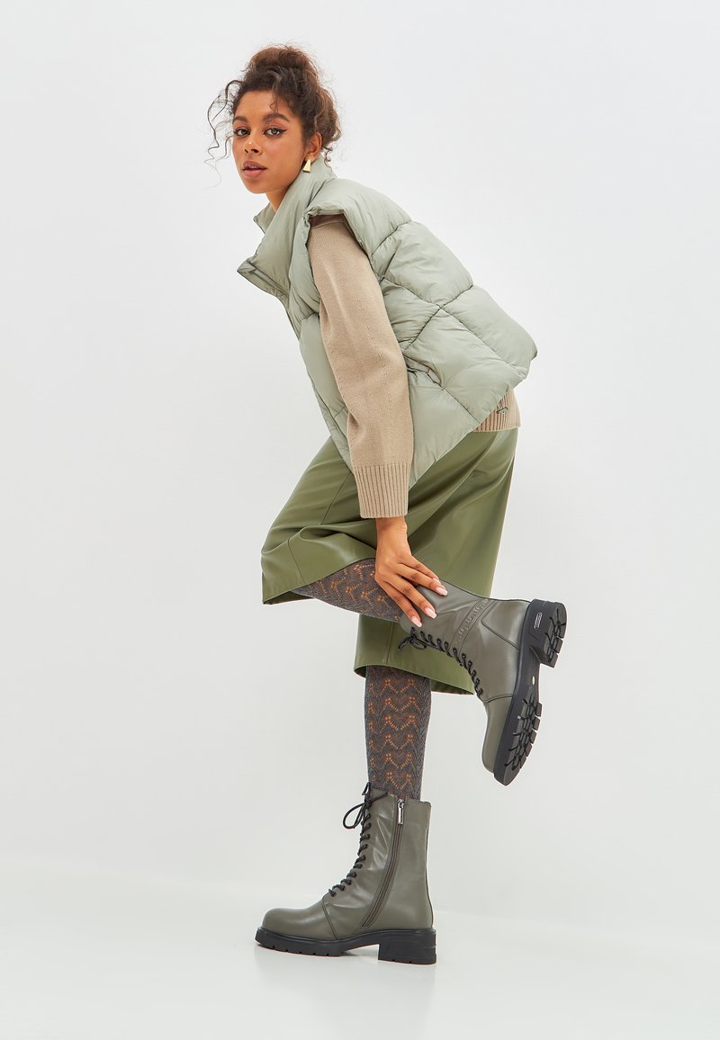 Dikke lichtgroene vest, beige trui, olijfgroene rok, patroontekenten panty’s en grijze veterschoenen met een stevige zool, voor een effen achtergrond.