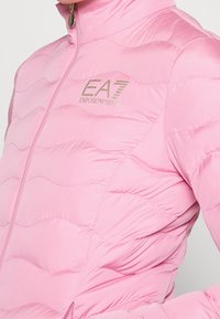 Světle růžová péřová bunda s texturou ve vlnkovém vzoru, zapínáním na zip po celé délce a zlatým logem EA7 Emporio Armani na oblasti hrudi.