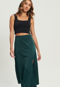 Schwarzes, kurzes Tanktop kombiniert mit einem dunkelgrünen Wickelrock mit seitlichem Schlitz, glattem Stoff und kantigem Design.