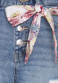 Shorts de mezclilla con botones de presión plateados, lavado azul claro y un detalle de lazo con estampado floral en la cintura. Incluye textura desgastada.