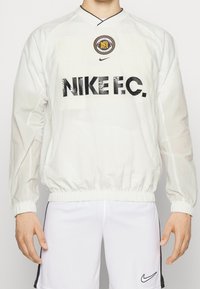 Biała lekka kurtka z okrągłym logo na lewej piersi, z napisem "NIKE F.C." dużymi czarnymi literami, elastycznymi mankietami i pasem, o gładkiej teksturze.