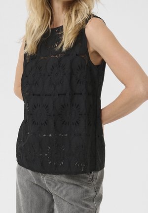 Vrouw die een mouwloos zwart kanten top met bloemmotieven en een grijze gestreepte broek draagt, staand met één arm achter haar rug.