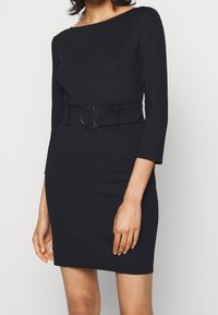 Schwarzes, figurnahes Kleid mit Dreiviertelärmeln, einem Boot-Ausschnitt und einem breiten Gürtel mit einer runden Metall-Schließe. Glattes, dehnbares Material.