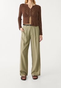 Bruine gebreide cardigan met een V-hals en knopen, gecombineerd met losse, olijfgroene broeken. Model draagt bruine sandalen en staat voor een effen achtergrond.