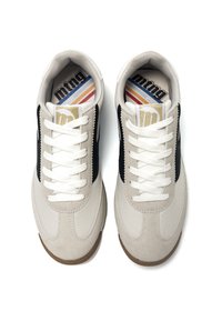 Sneaker mit einem beigefarbenen Textil-Obermaterial und Wildlederakzenten, schwarzen Seitenpaneelen, weißen Schnürsenkeln und einer kontrastierenden braunen Gummisohle.