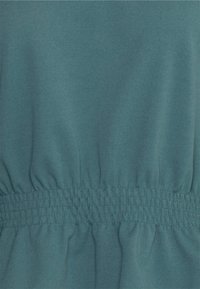 Teal fabric with a smooth texture features an elasticated waistband, creating gathers for a relaxed fit. Simple and uniform design.