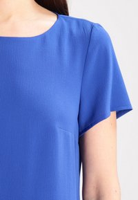 Krátký rukáv blůza v jasně modré barvě, s texturovanou tkaninou, kulatým výstřihem a malou kapsou na přední levé straně.
