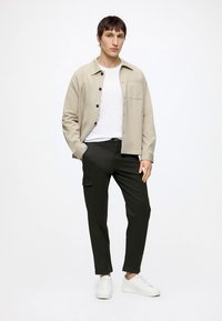 Giovane uomo in piedi con le mani in tasca, indossa una giacca camicia beige con bottoni, una maglietta bianca, pantaloni cargo neri e scarpe da ginnastica bianche.