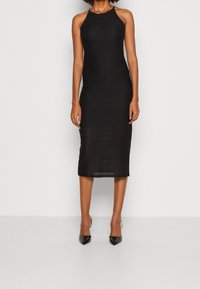 Vestido midi negro con escote halter, fabricado en tejido texturizado, de silueta ajustada y con abertura lateral. Llevado con tacones negros de punta.