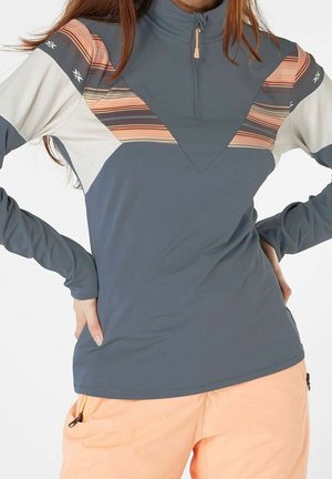 Pull gris à manches longues avec des accents pêche, blanc et rayés, avec une fermeture éclair au quart, porté avec un pantalon pêche, modèle mains sur les hanches.