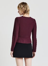 Maglione lavorato a maglia color bordeaux con maniche lunghe a costine e dettagli testurizzati, collo ondulato e orlo plissettato, indossato sopra una gonna nera.