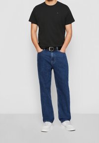 Man in een zwart t-shirt, blauwe jeans, zwarte riem en witte sneakers, staand met zijn handen in zijn zakken.